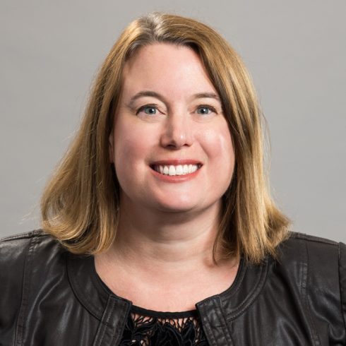 A woman with shoulder-length blonde hair smiles at the camera, wearing a black leather jacket and a black top, in front of a plain grey background.
