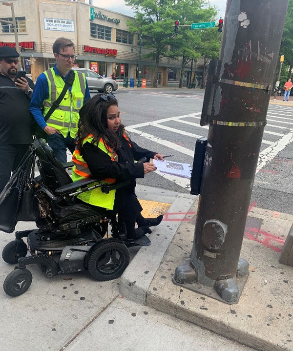 This pedestrian is unable to reach an Accessible Pedestrian Signal in Downtown Silver Spring because the push button is located on a raised surface that her wheelchair cannot navigate.