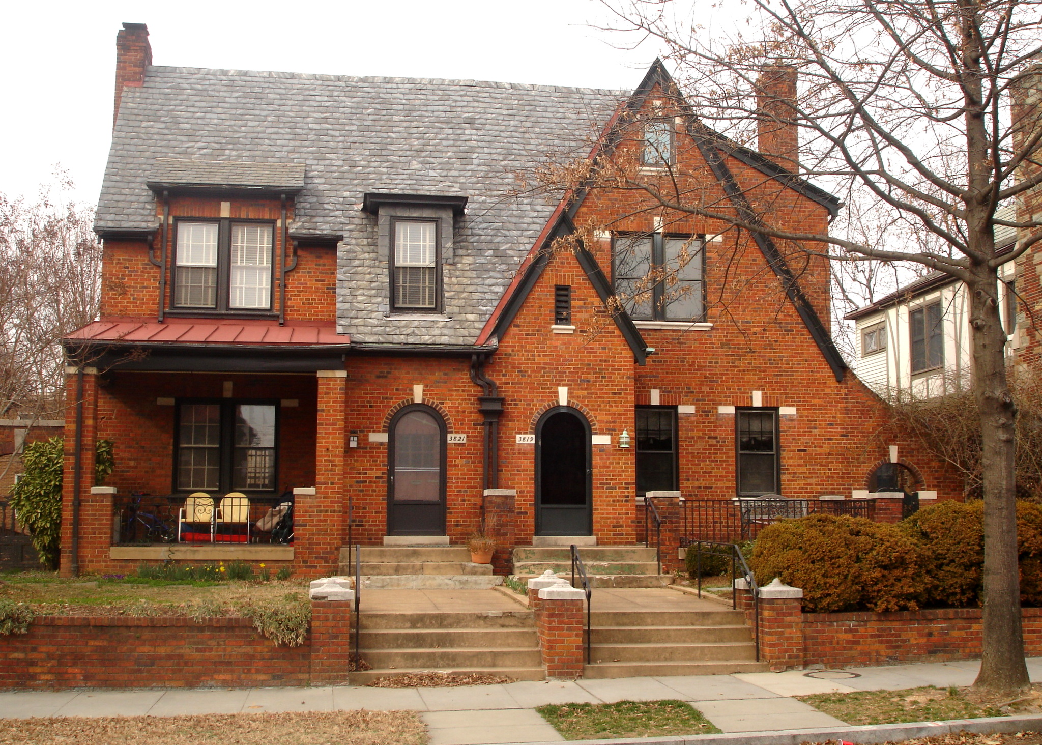 A brick duplex with a gabled roof, red awning over the porch, two front doors, and a small front yard with bare trees.