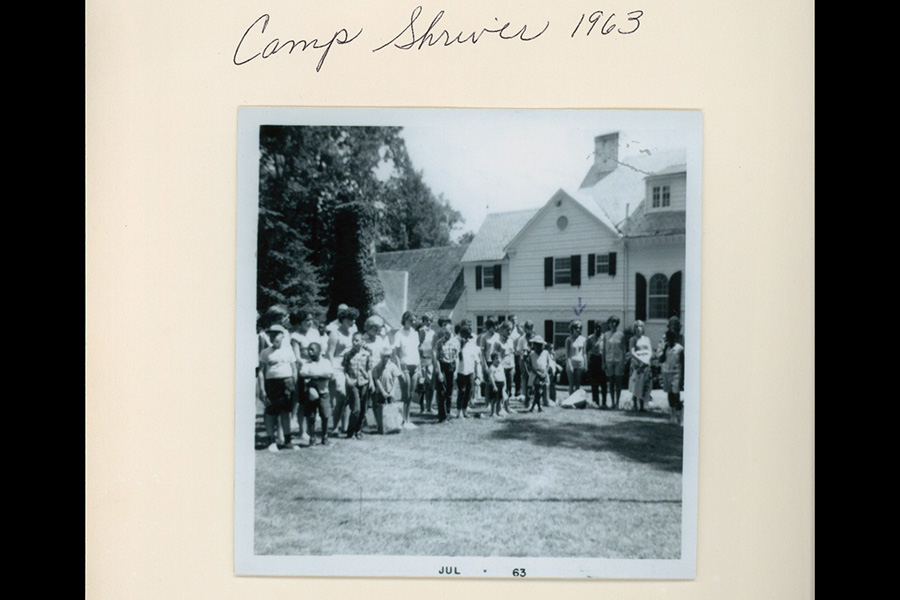 Black-and-white photo labeled "Camp Shriver 1963" at the top. A group of children and adults stands in front of a large house on a grassy lawn. Trees are visible in the background. Photo dated July 1963 at the bottom.