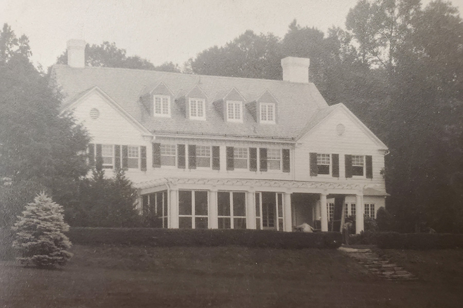 A large, classic two-story colonial-style house with a gabled roof, multiple chimneys, and a front porch surrounded by trees and shrubs. The black-and-white photograph gives it a vintage appearance, suggesting an early 20th-century setting.