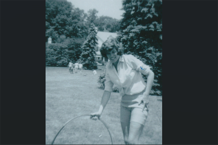 A woman in a short-sleeved shirt and shorts is smiling while holding a hula hoop on a grassy lawn. Trees and a house are visible in the background on a sunny day.