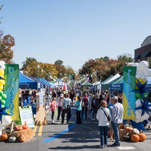 A lively outdoor street festival with people exploring vendor booths under blue and green tents. The entrance is decorated with balloons and signs reading "Damascus" and "Placemaking." Trees with autumn leaves line the street, and pumpkins are displayed.