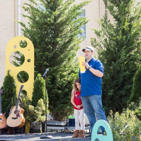 A man in a blue shirt and cap holds a yellow paper while speaking into a microphone on an outdoor stage. A young girl stands beside him, partially hidden. A guitar rests on the stage nearby, with decorative panels and greenery in the background.