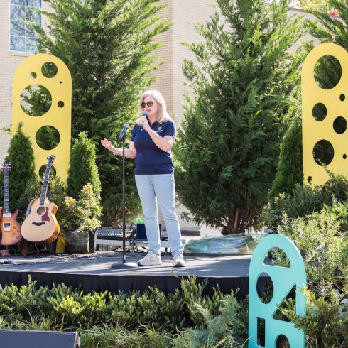 A woman in sunglasses speaks into a microphone on an outdoor stage. She stands between two yellow and green sculptures with cutout designs, surrounded by trees and musical instruments.