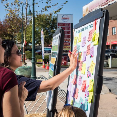 People reading and adding sticky notes to a large display board outdoors. The board is covered with colorful notes, and a gas station can be seen in the background on a sunny day.