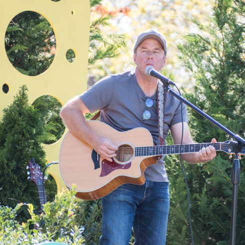 A man in a gray t-shirt and cap plays an acoustic guitar while singing into a microphone outdoors. He stands in front of decorative cutouts and greenery. Sunglasses hang on his shirt as he performs.