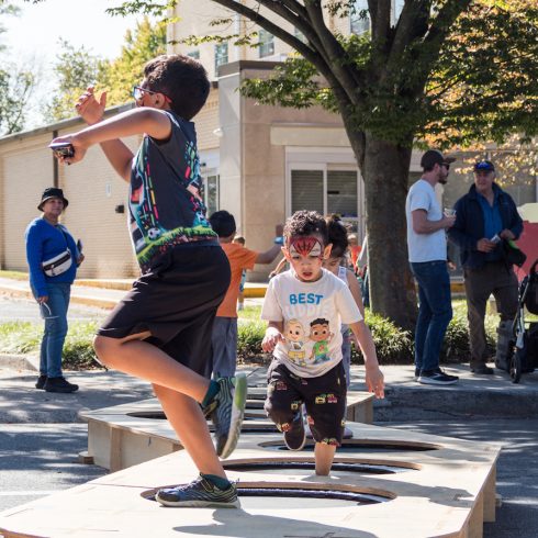 Children are energetically jumping on a wooden platform in an outdoor area with adults watching. One child wears a shirt with cartoon characters. Trees and a building are visible in the background, along with several spectators.