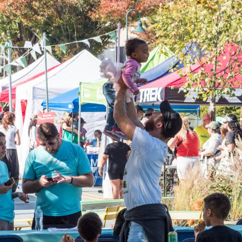 A man lifts a smiling toddler into the air at an outdoor festival. Colorful tents and people are in the background. Tables with seated adults and children are in the foreground, creating a lively family-friendly atmosphere.