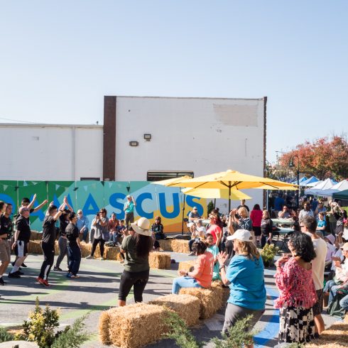 A lively outdoor event with people enjoying music and performances. Attendees sit on hay bales under yellow umbrellas, while a group of dancers performs. Tents and colorful letters spelling "ACUS" are in the background, with a clear blue sky above.