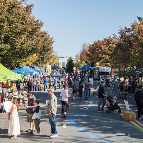 A lively outdoor street festival with people walking and visiting food trucks and tents. Trees with autumn foliage line the street under a clear blue sky. Families and groups are enjoying the sunny day.