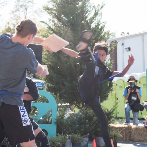 A martial artist performs a high kick to break a board held by another person outdoors. People watch in the background, and a photographer captures the moment.