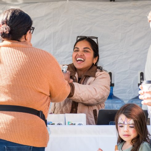 A smiling woman hands an item to another woman in front of a booth. A young girl with face paint stands nearby, holding a cup. Everyone appears to be enjoying the interaction outdoors.