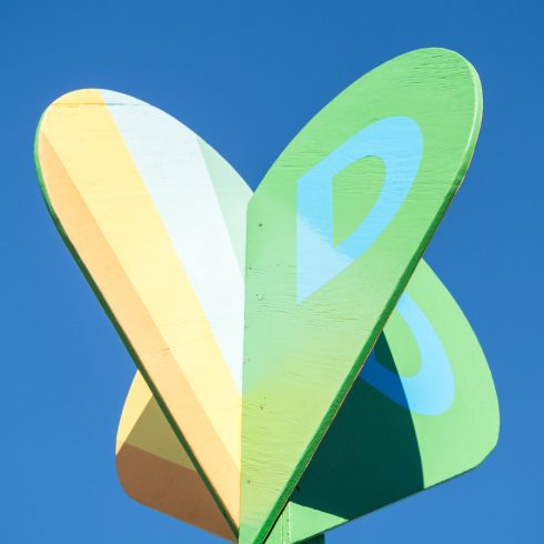 Colorful wooden structure with heart-shaped panels in yellow, green, and light blue, set against a clear blue sky. Each panel has a different color and design, creating a playful and artistic look.