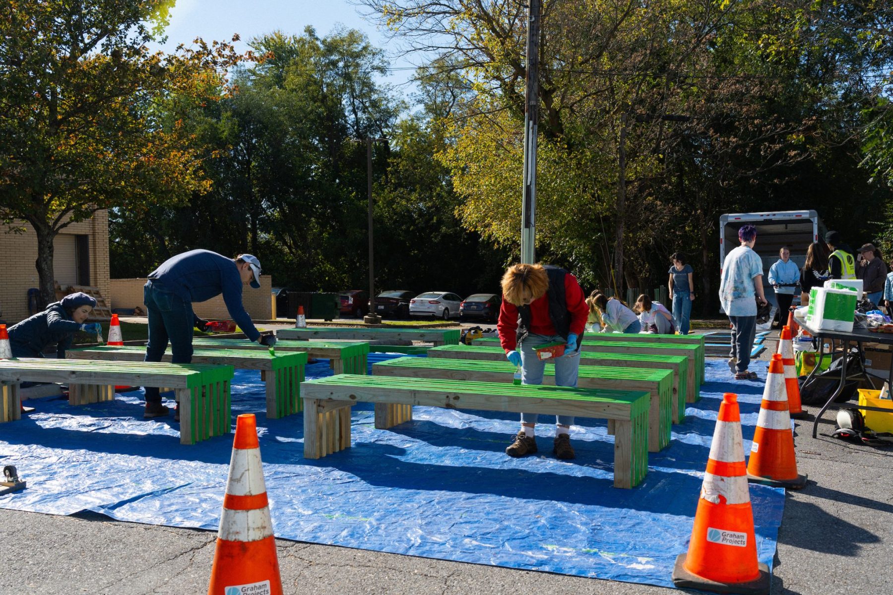 People painting benches on a blue tarp outdoors. Orange traffic cones are placed around the area. The scene is under sunny weather, surrounded by trees and a few parked cars.