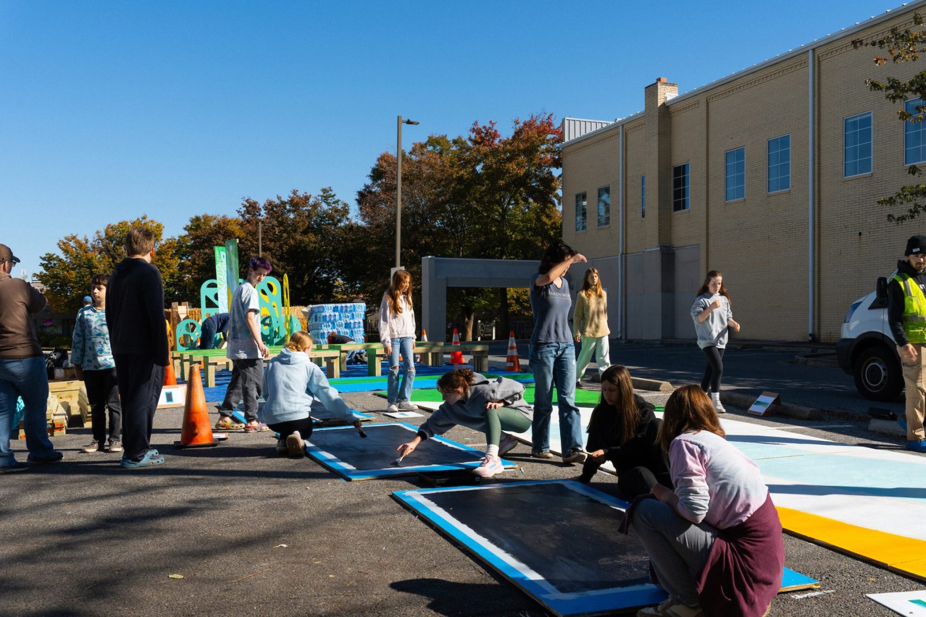 People painting large boards on the ground outside a yellow brick building. Traffic cones and art supplies are scattered around. It's a sunny day, and autumn trees are visible in the background.