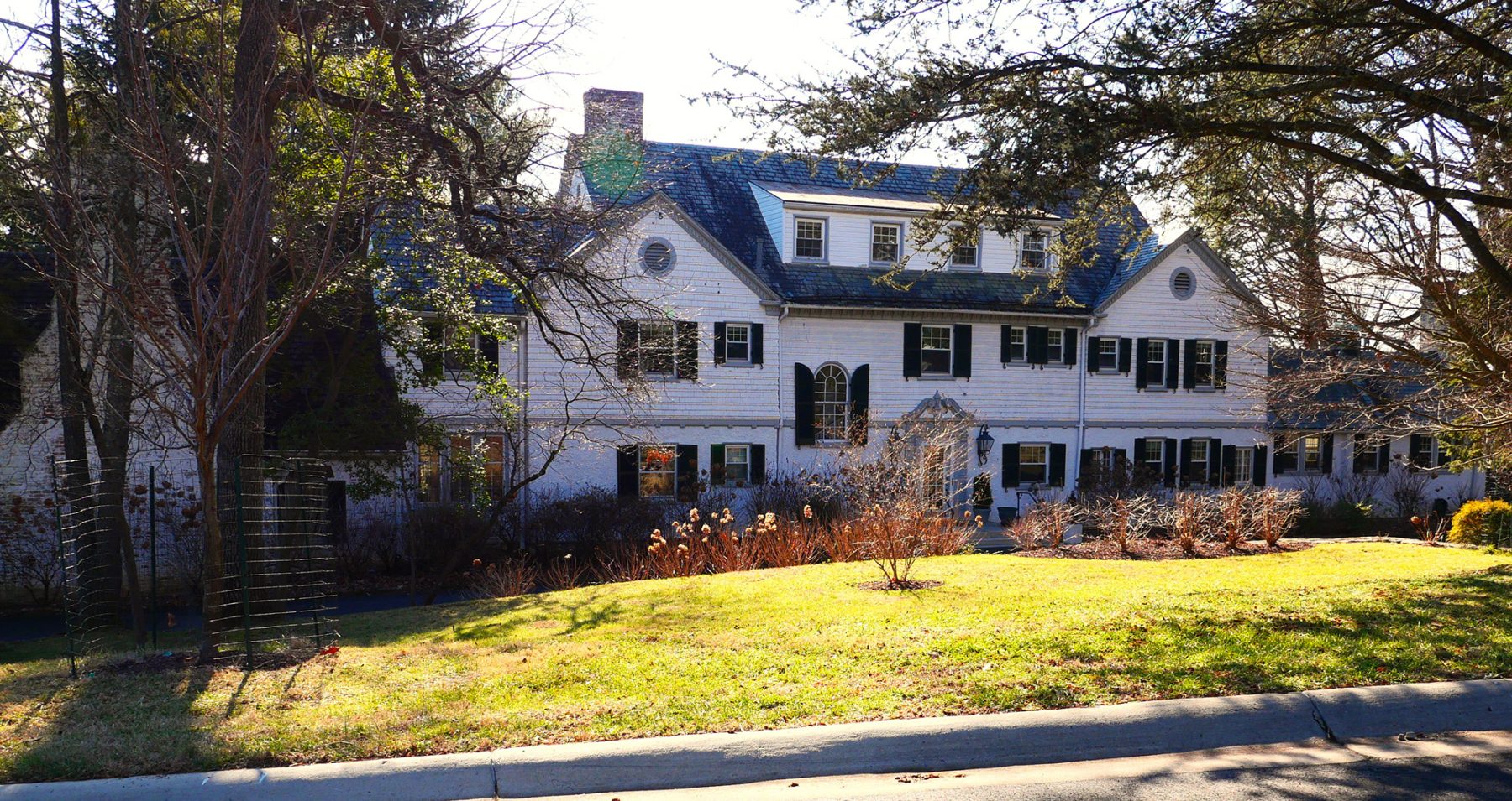 A large, two-story white house with dark green shutters sits surrounded by bare trees and shrubs. The sun casts bright light on the house and the lawn in front, with a clear blue sky above.