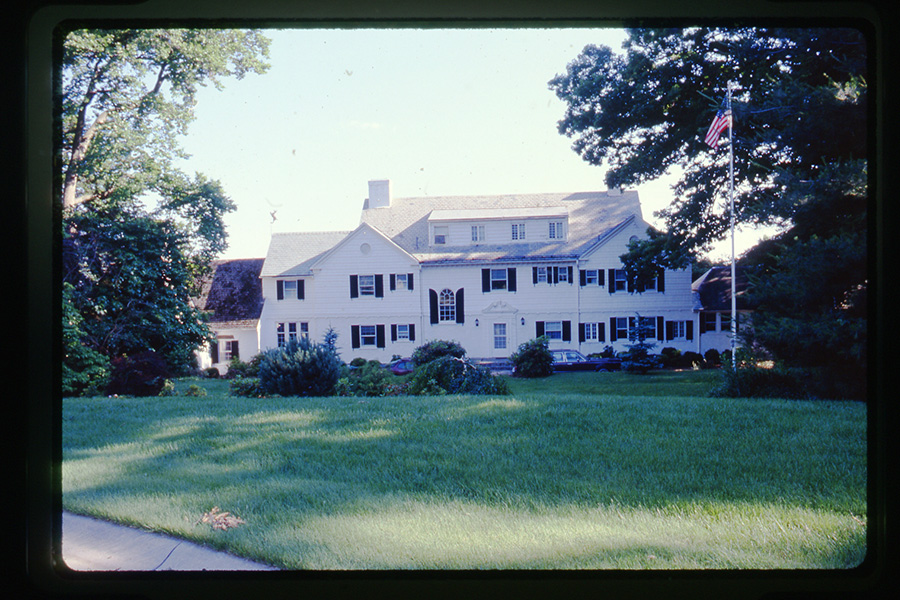 A large, two-story white colonial house with black shutters is surrounded by lush green lawns and trees. An American flag flies on a tall flagpole in the front yard under a clear blue sky.