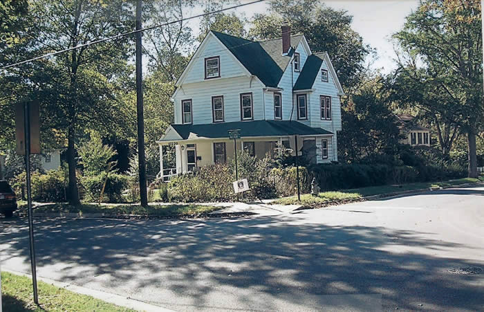 A two-story house with green roof sits at a corner surrounded by trees and shrubs. A small sign is visible near the street.