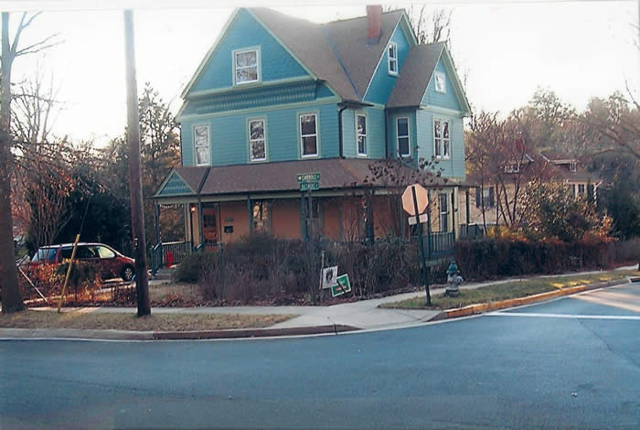 A large, blue, two-story house with an attic and a wraparound porch is situated at a street corner, surrounded by leafless trees and a parked car in the background.