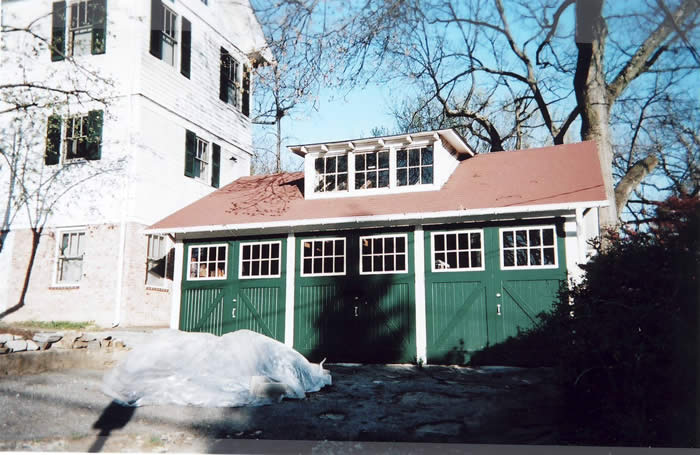 A three-car garage with green doors and a red roof next to a white house. Plastic sheeting is on the ground in front.