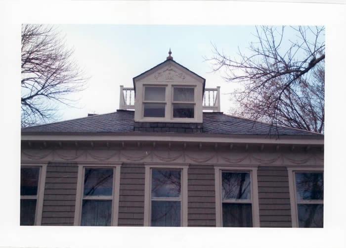 A house roof with an attic window and small balcony, flanked by bare tree branches under a clear sky.