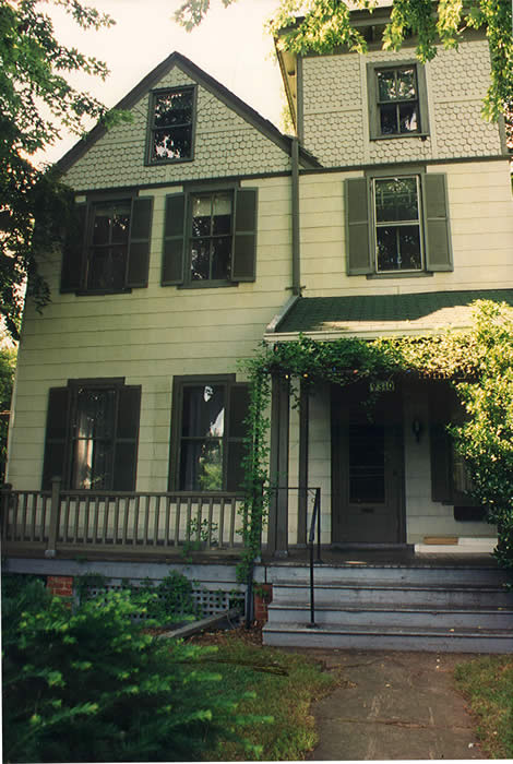 A two-story house with pale yellow siding and green shutters, partially covered by greenery. A porch with a railing is at the front.