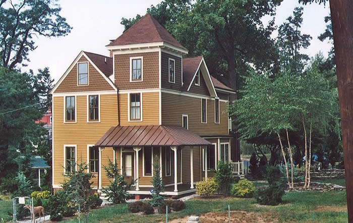 A large, multi-story Victorian-style house with brown siding, white trim, and a steep roof. Surrounded by trees and shrubs, it sits on a landscaped lot.