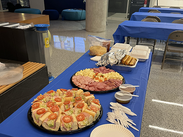 A table with assorted sandwiches, cheese, and meats on platters, accompanied by disposable plates, utensils, and condiments, set up for a gathering.