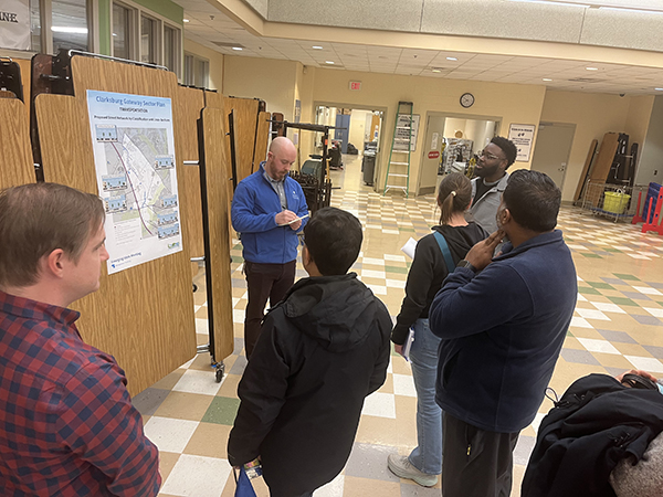 A group of people stand in a hallway listening to a person speaking in front of a presentation board.