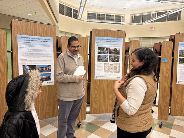 Two adults and a child interact in front of informational display boards titled "Clarksburg Gateway Sector Plan" in a community meeting space.