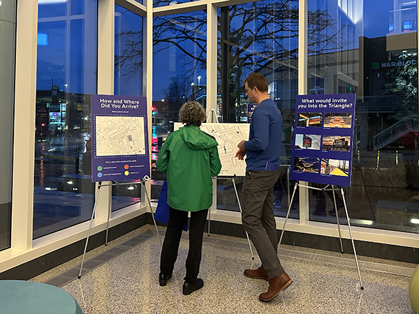 Two people stand in front of informational posters on easels inside a building with large windows, discussing maps and images related to urban planning.