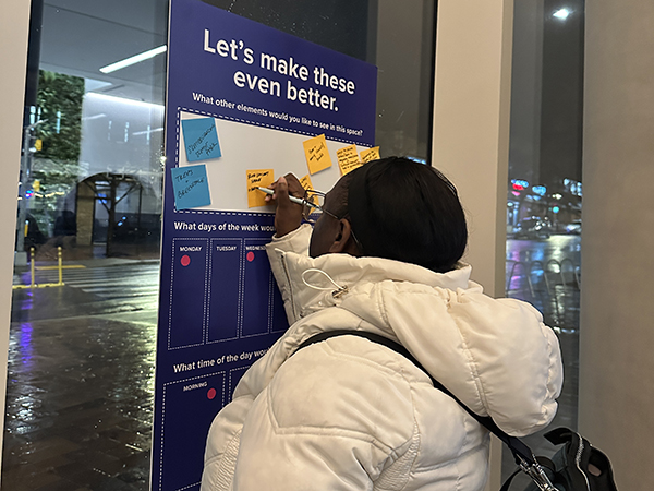 A person in a white coat writes on a sticky note attached to a suggestion board indoors, titled "Let's make these even better.