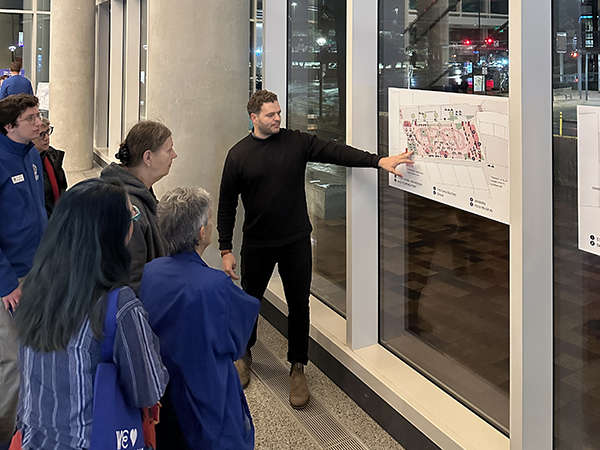 A person in a black sweater presents a map to a group of people inside a building with large windows.