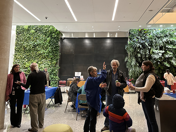 People gather indoors near a vertical garden wall. Some are standing and talking, while others are seated at tables.