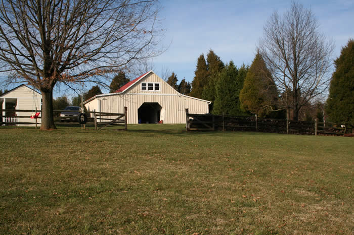 A white barn with a red roof stands in a grassy field, surrounded by trees and a wooden fence. A smaller building and a parked vehicle are nearby.