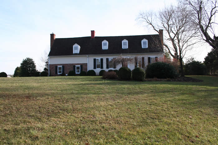 Large, two-story colonial house with a dark roof, white and brick exterior, and multiple windows. Surrounded by grass and bushes, with bare trees in the background under a clear sky.