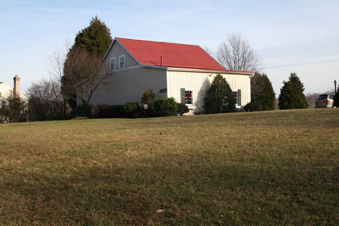 A building with a red roof and white siding is surrounded by trees and bushes, set on a grassy lawn under a clear sky.