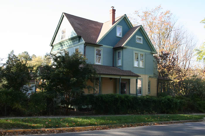 Two-story green and yellow house with a chimney, surrounded by trees, viewed from the street.