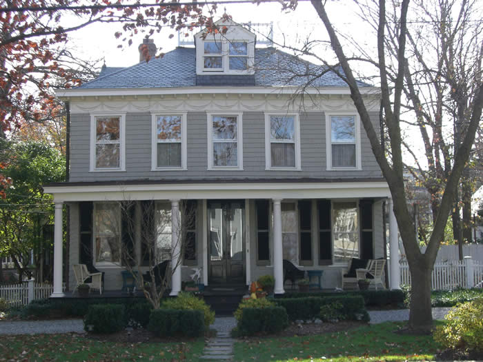 Two-story gray house with a large porch, white columns, and black shutters. Surrounded by trees and bushes with a small path leading to the front steps.