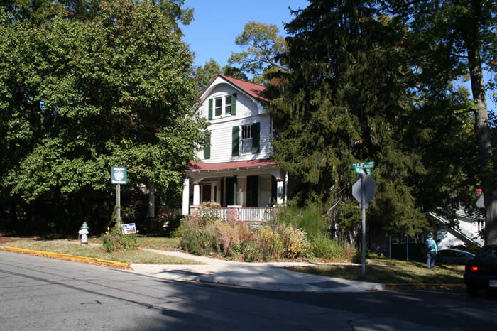 A two-story house with a porch and red roof, surrounded by trees and shrubs, is located at a street intersection.