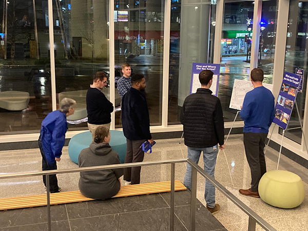 A group of people stand indoors, observing posters displayed on easels. A city street is visible through the large windows in the background.