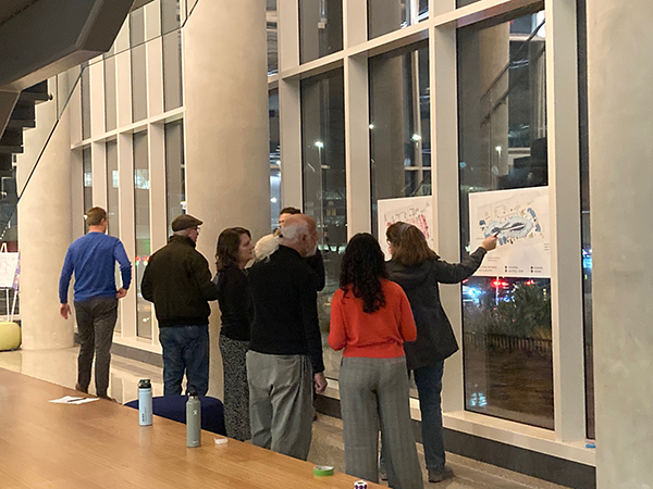 A group of people stands by large windows, discussing and pointing at two posters displayed on the glass.