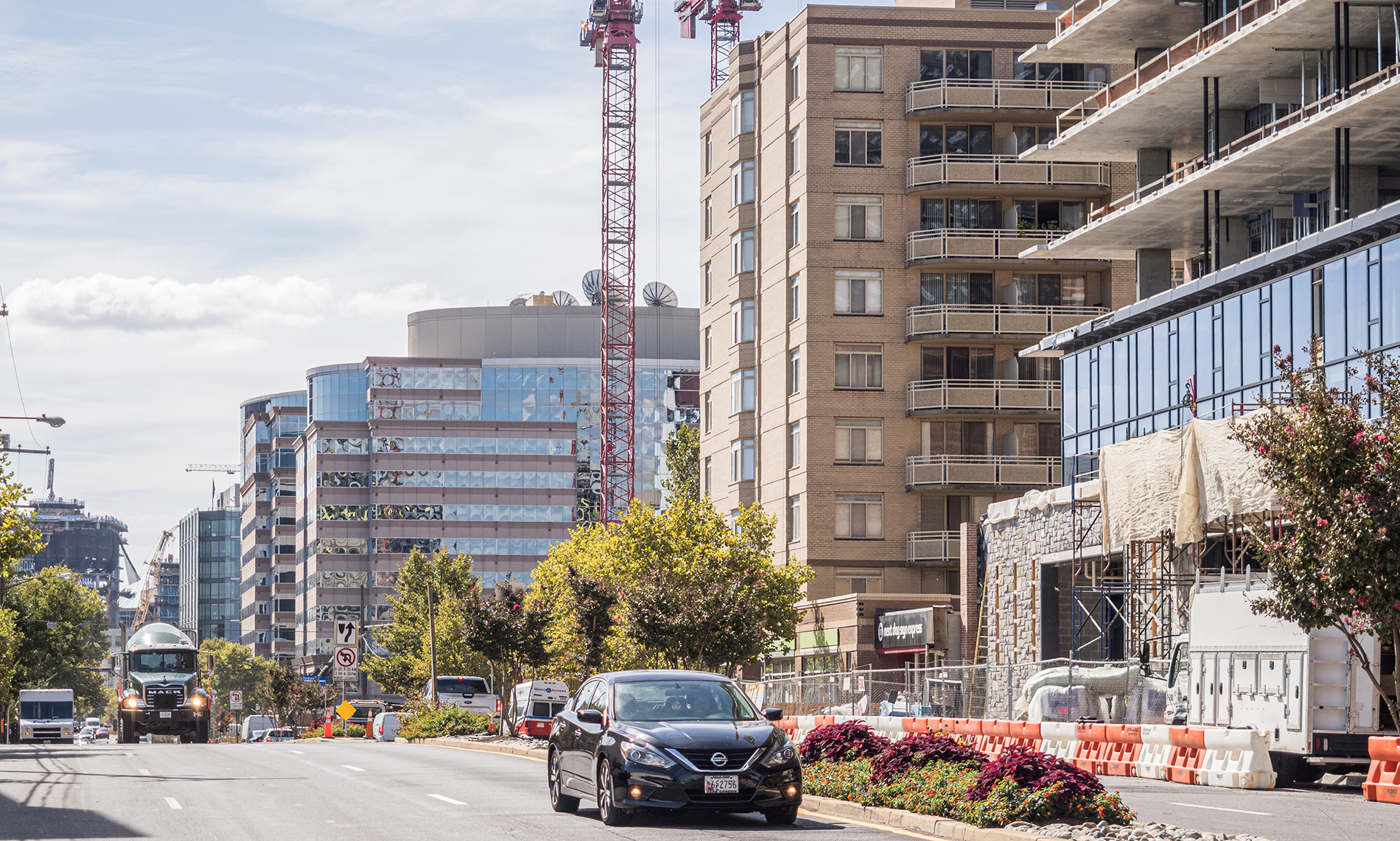 Street view of an urban area with modern buildings, a construction site, and cars on the road. Two cranes rise above the buildings under a partly cloudy sky.