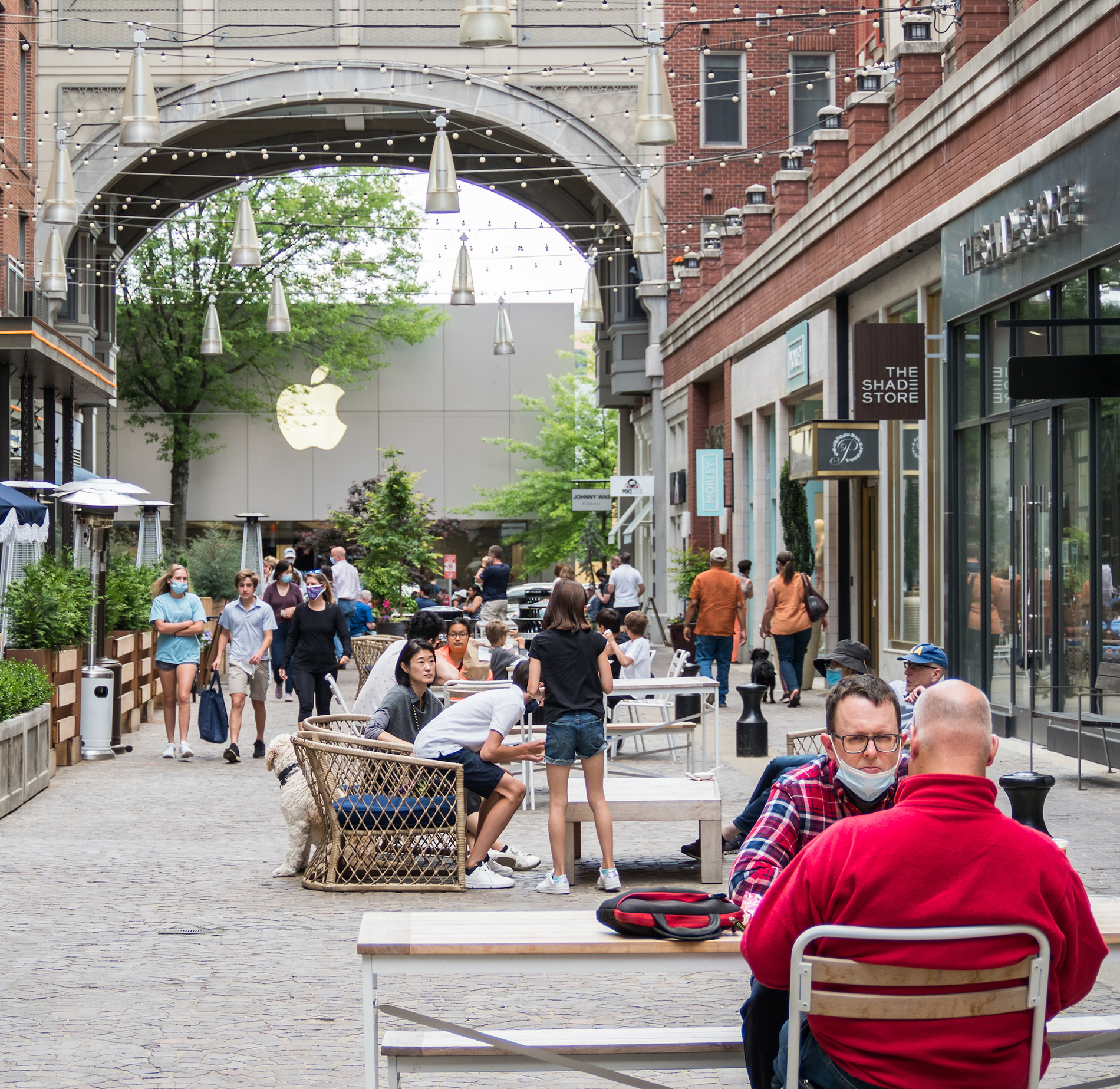 Outdoor shopping area with people walking and sitting at tables. Shops line the sides, and an Apple store is visible in the background under string lights.