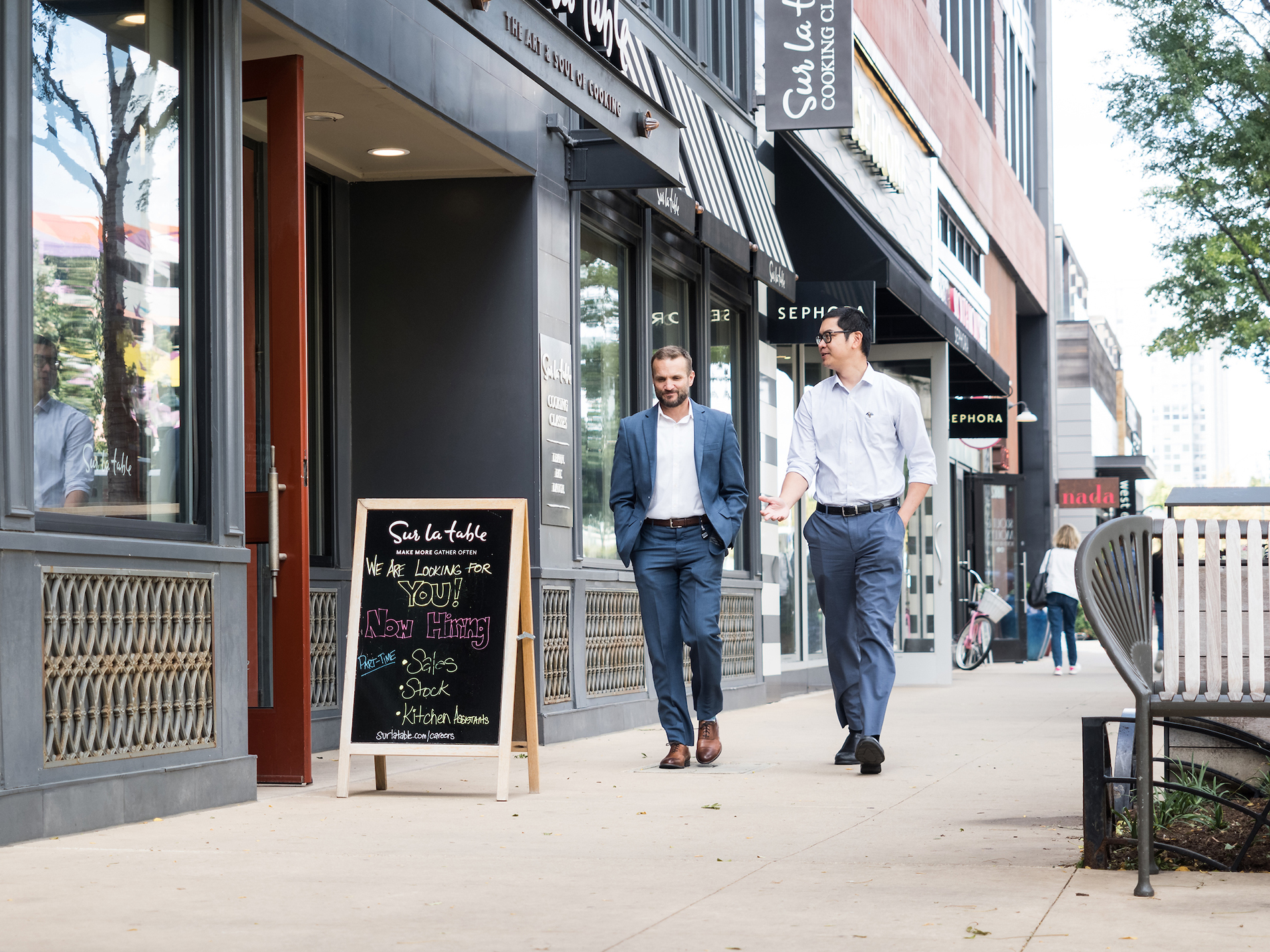 Two men in business attire walk down a sidewalk past storefronts with a chalkboard sign outside a restaurant.