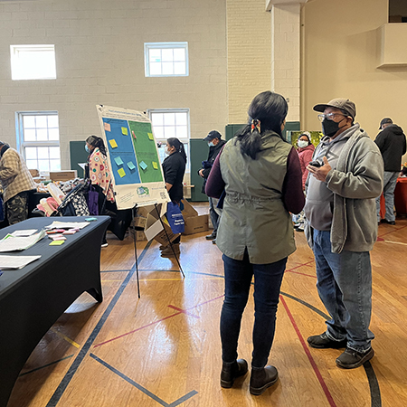 People in a gymnasium engage in conversation around a display board with sticky notes, some wearing masks.