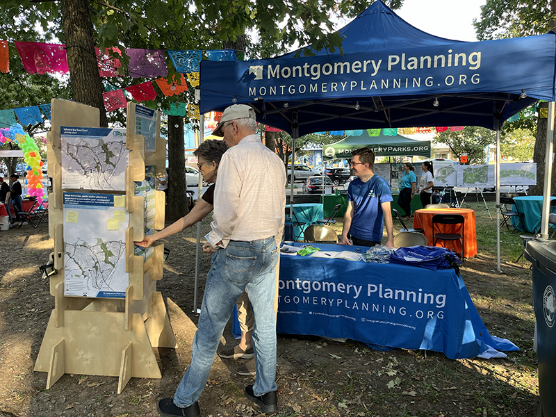 People view maps at a Montgomery Planning booth with informational displays and a tent at an outdoor event.