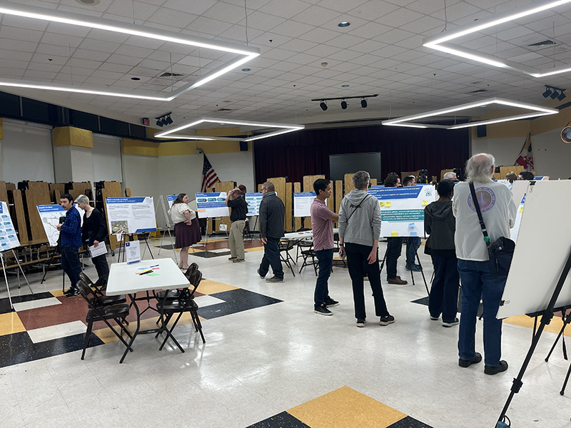 People observing various informational posters displayed on easels in a large indoor room with checkered flooring and ceiling lights.