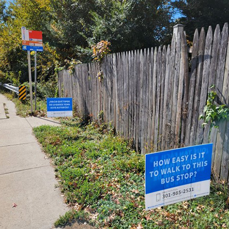 Wooden fence lining a sidewalk with blue signs asking about ease of access to a bus stop. Bus stop sign and contact number visible.